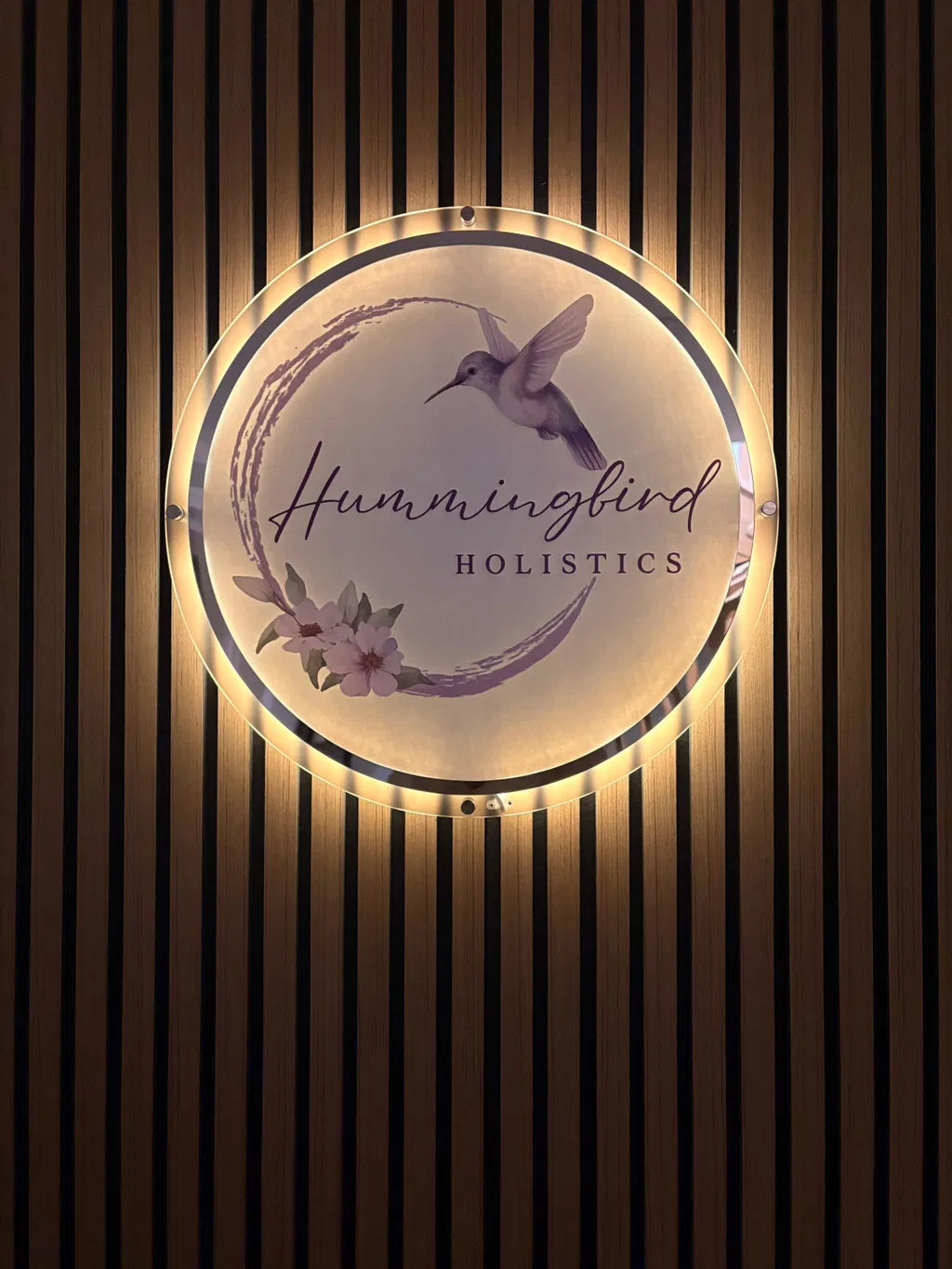 A circular illuminated sign for Hummingbird Holistics mounted on a dark wood slatted wall. The sign features a hummingbird illustration, delicate floral details, and the business name in elegant script and serif fonts, glowing warmly against the background.