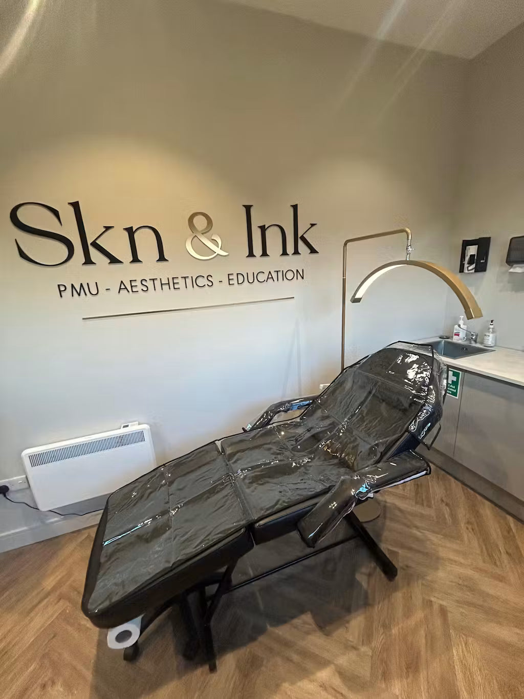 Interno di uno studio di tatuaggi con un lettino per i trattamenti e un'insegna in acrilico personalizzata del marchio "Skn & Ink" appesa alla parete.